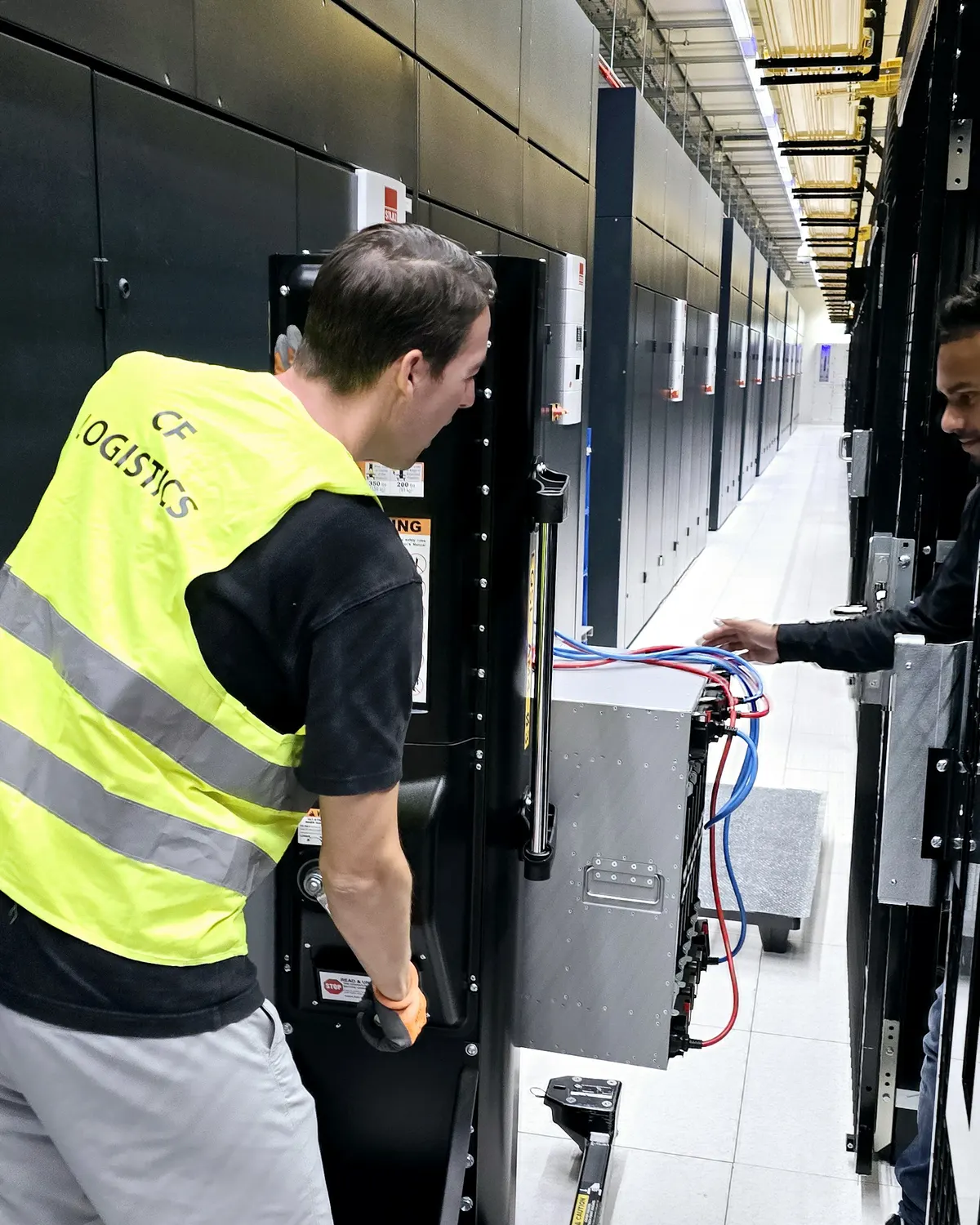 A worker in a hi-vis vest operating a server lift beside a rack in a data hall while a colleague frees the cables.